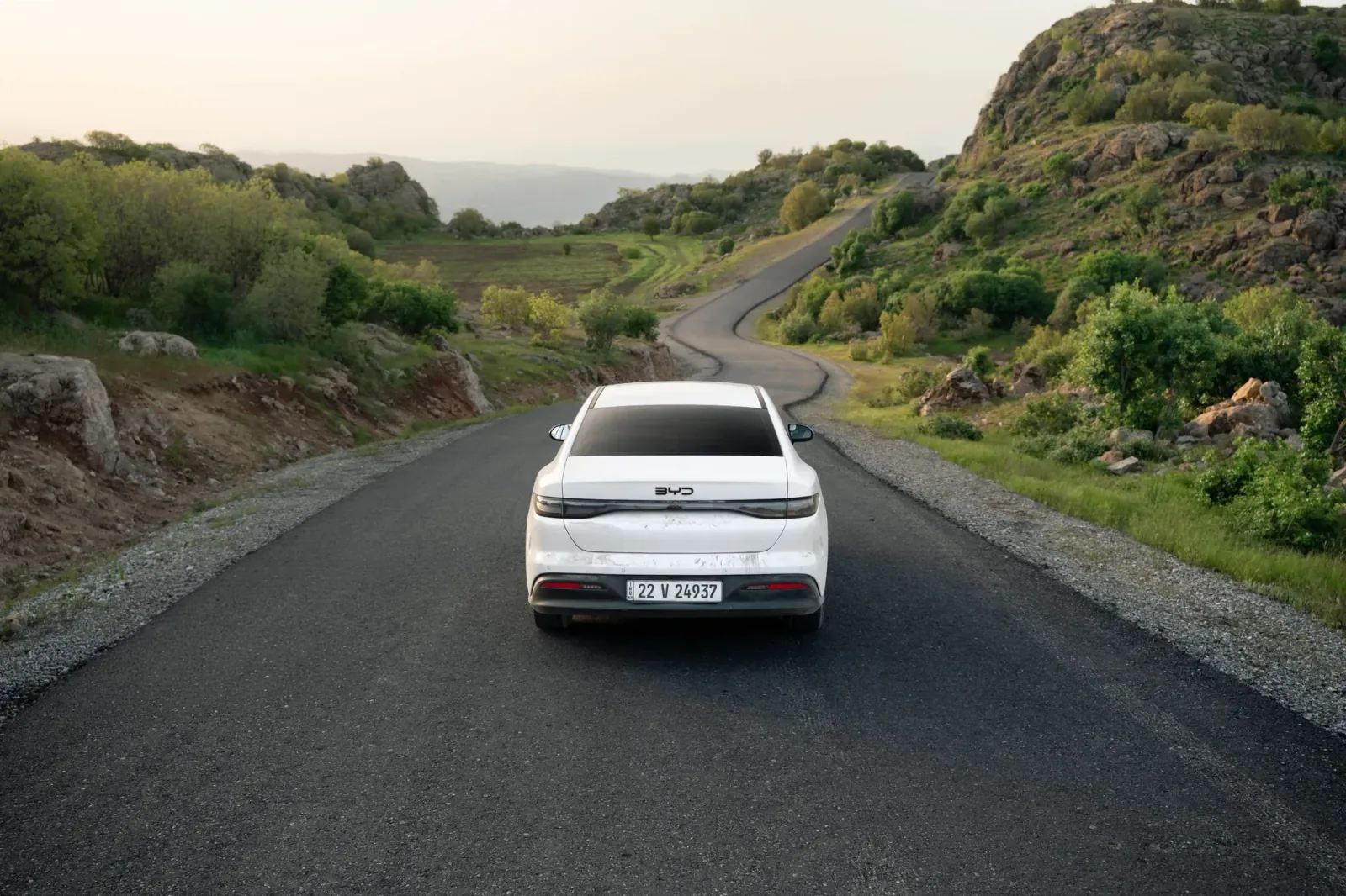A white car travels down a winding road in the lush hills of Shaqlawa, Iraq, showcasing serene scenery.
