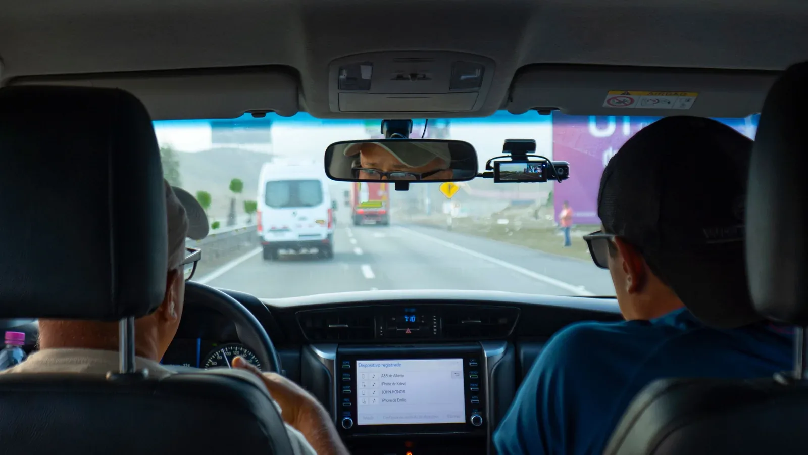 Interior of a car with two people driving on a highway, focusing on dashboard and rear view mirror.
